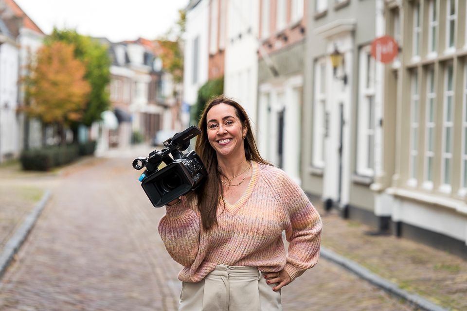 Vrouw met een videocamera lacht op een straat met kinderkopjes. Beige trui, gebouwen op de achtergrond.