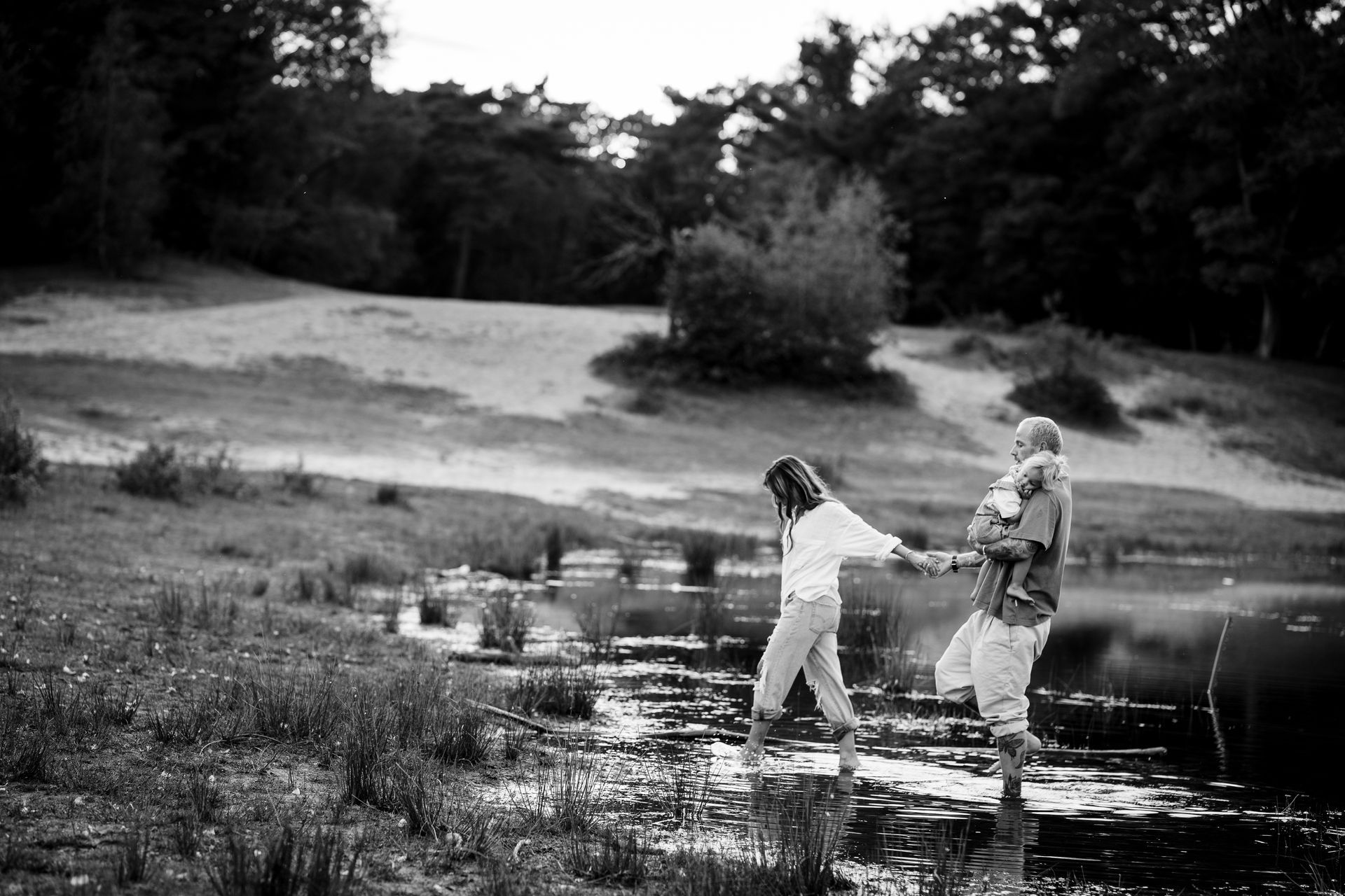 Een stel waadt door het water met een kind op de arm. Het landschap bestaat uit bomen, een meer en glooiende grasvelden.