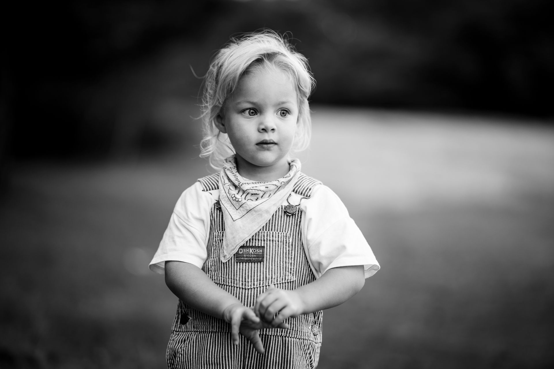 Jong kind met blond haar, gekleed in een tuinbroek en een bandana, kijkt buiten beeld in een buitenomgeving.