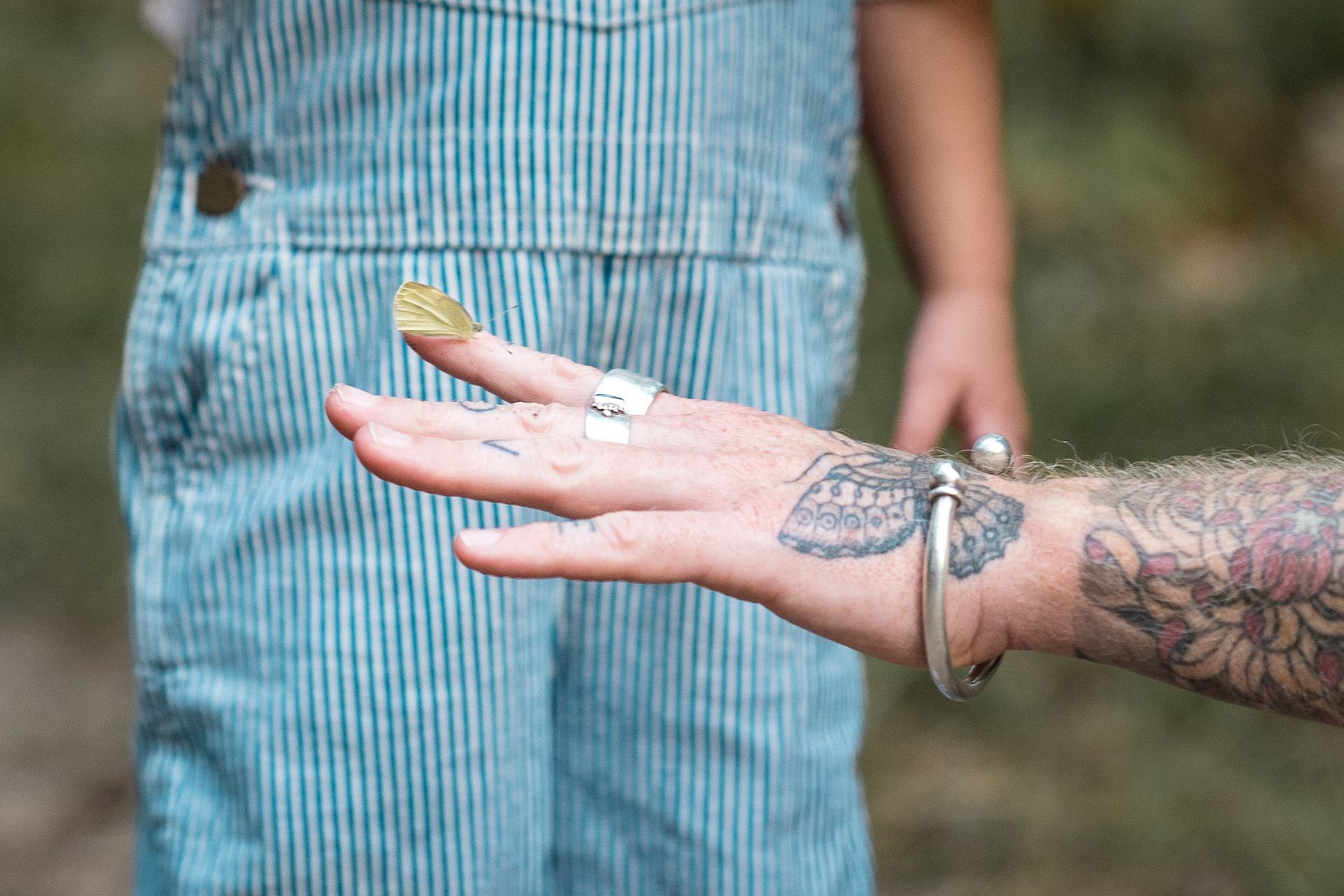 Hand met tatoeages die een geel blad vasthoudt; kind in blauwe overall op de achtergrond.