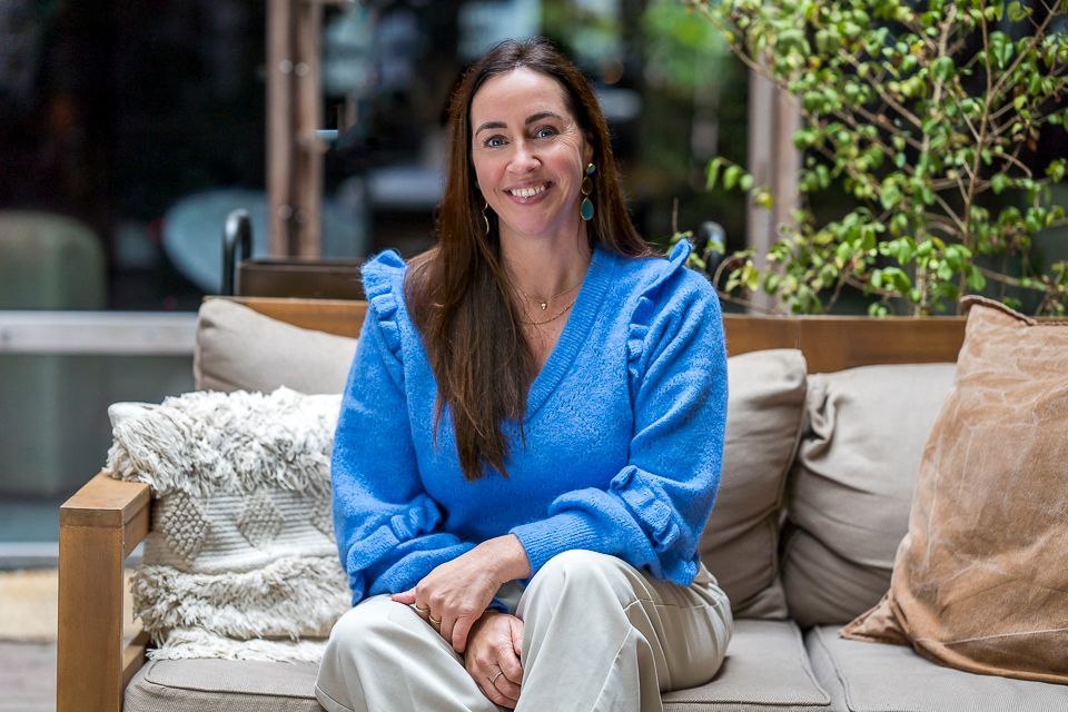 Vrouw zit op een bank, gekleed in een blauwe trui en beige broek, en lacht.