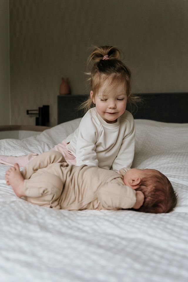 Een kind kijkt neer op een pasgeboren baby op een bed. De baby is ingewikkeld in een doek, waardoor het gezichtje bedekt is.