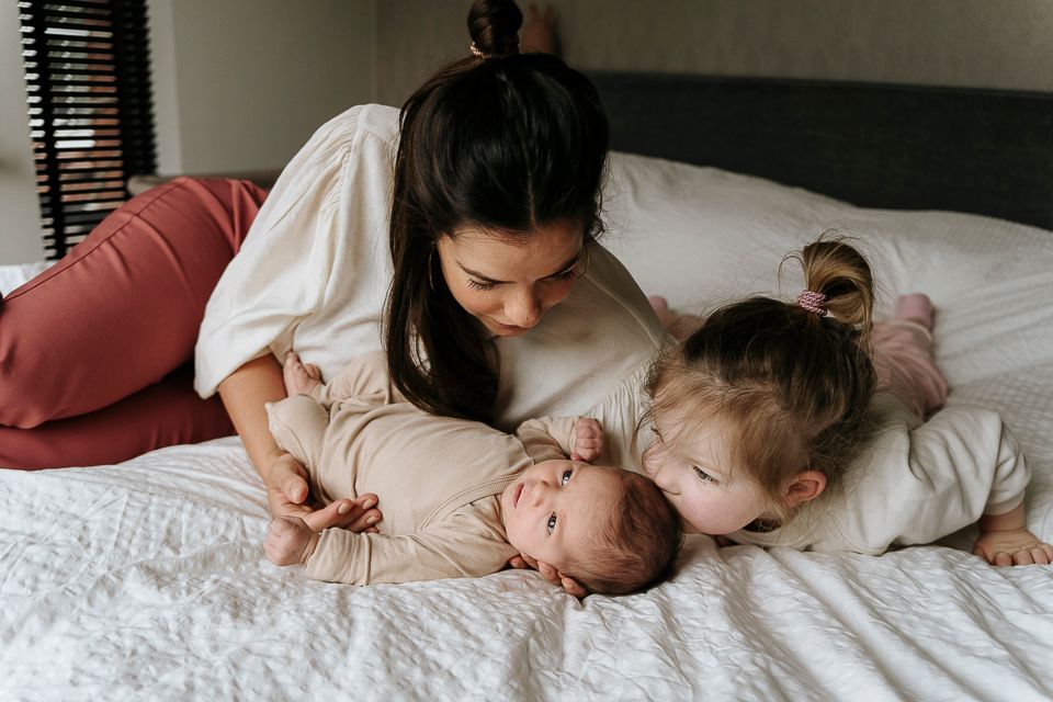 Vrouw en twee jonge kinderen op een bed. De vrouw kijkt naar de baby in haar armen terwijl een van de kinderen naar haar toe buigt.