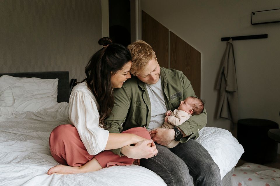 Ouders wiegen hun pasgeboren baby in bed; ze kijken liefdevol naar de baby. Slaapkamer, neutrale tinten.