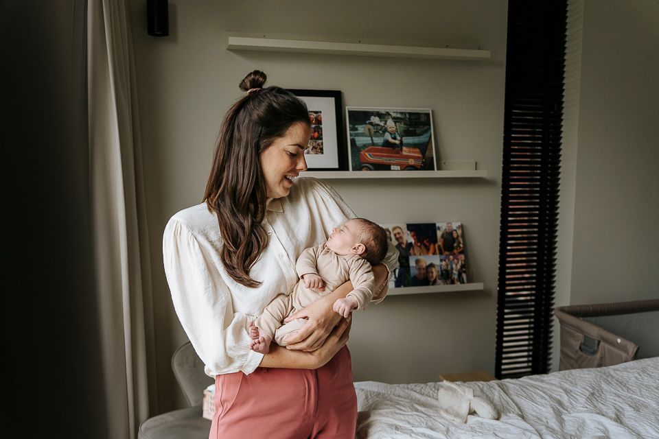 Vrouw met baby in haar armen, lachend, in een lichte kamer. Planken met foto's zijn zichtbaar.
