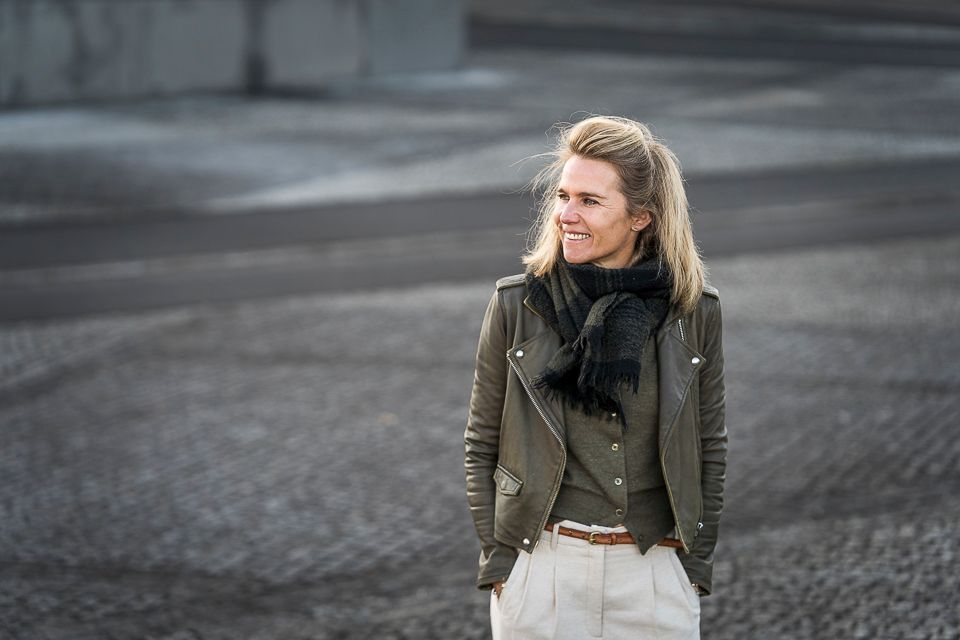 Een lachende vrouw staat buiten, gekleed in een groene jas, een zwarte sjaal en een witte broek; haar handen zitten in haar zakken.
