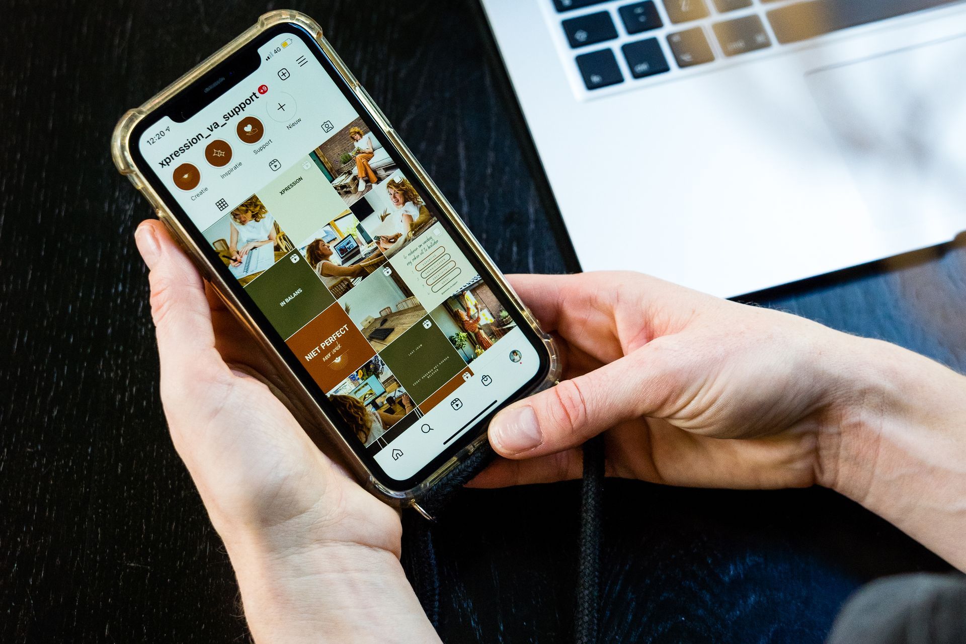 Handen houden een smartphone vast waarop een Instagram-feed te zien is. De telefoon is verbonden met een laptop.