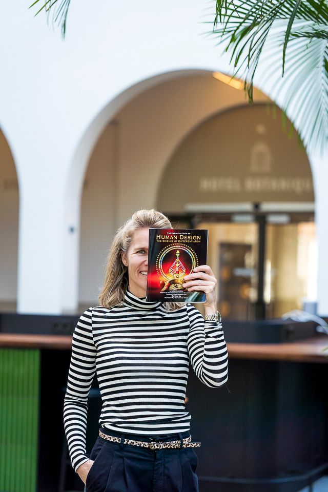 Vrouw met een boek voor haar gezicht. Wit-zwart gestreept shirt, zwarte broek. Buitenomgeving met bogen.