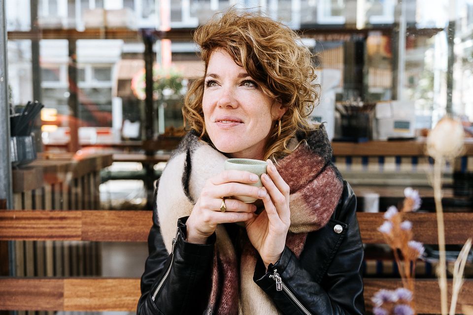 Vrouw met een kopje in haar hand, opzij kijkend, gekleed in een sjaal en jas, zit buiten.