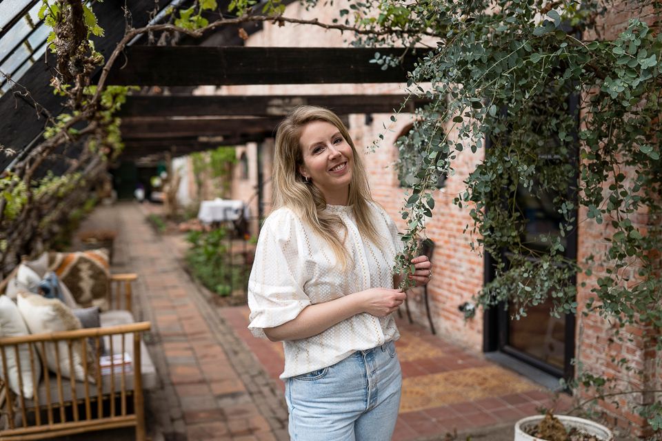 Een lachende vrouw houdt een plant vast in een tuin met bakstenen muren en een houten pergola.