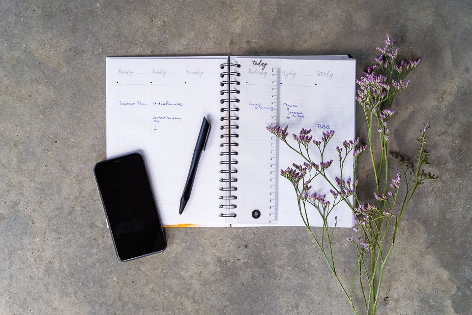 Open agenda met pen, smartphone en paarse bloemen op een grijze betonnen ondergrond.
