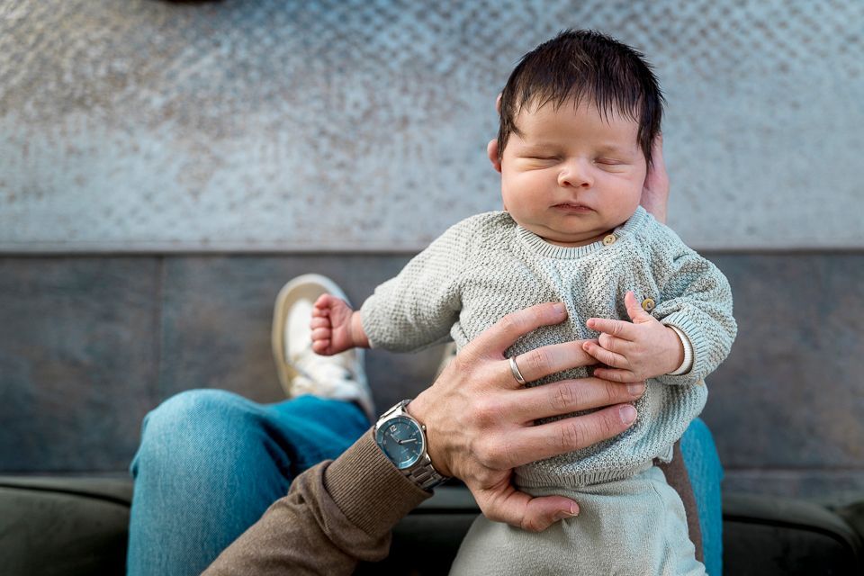 Een baby, met gesloten ogen en gekleed in een lichtgrijze trui, ligt rechtop op de schoot van iemand.