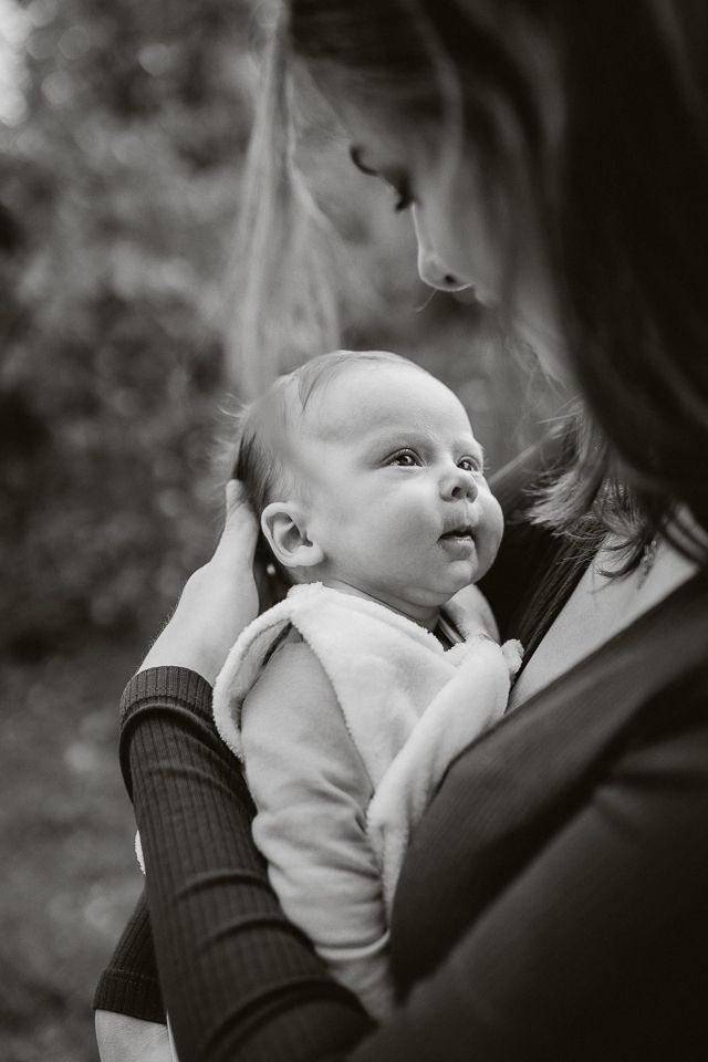 Vrouw houdt een baby vast en kijkt liefdevol naar beneden. De baby kijkt weg, ingewikkeld in een deken. Zwart-wit, buitenomgeving.