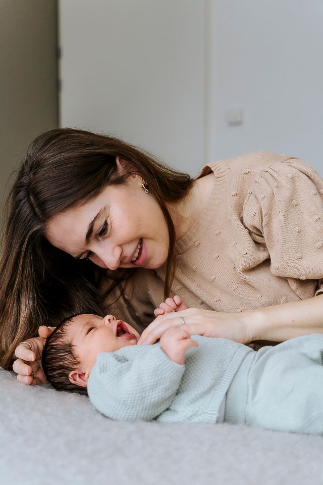 Een vrouw glimlacht naar een baby die op een bed ligt. De baby draagt ​​een blauw rompertje en kijkt omhoog.
