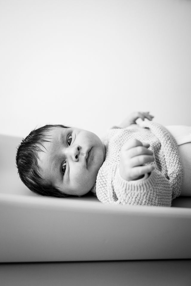 Pasgeboren baby ligt op een aankleedkussen en kijkt naar de camera. Houdt een gebreid voorwerp vast. Zwart-wit.