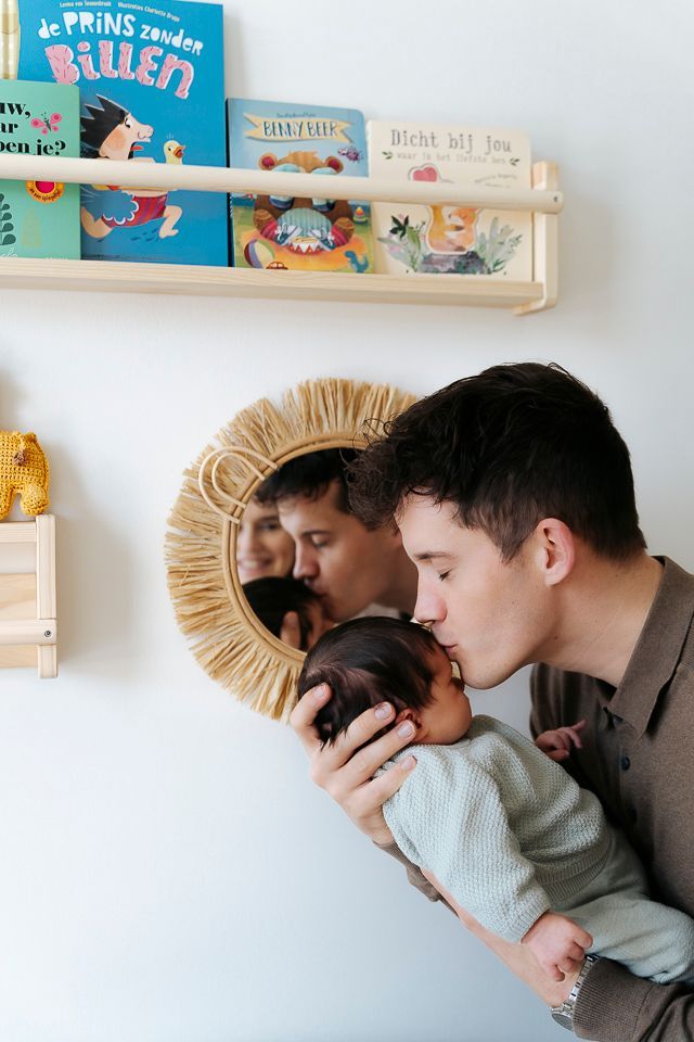 Een man kust een baby, weerspiegeld in een ronde spiegel. Witte muur, houten plank met boeken.