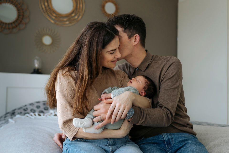 Ouders die hun pasgeboren baby vasthouden, vader die moeder op het hoofd kust, zittend op bed voor een wanddecoratie.