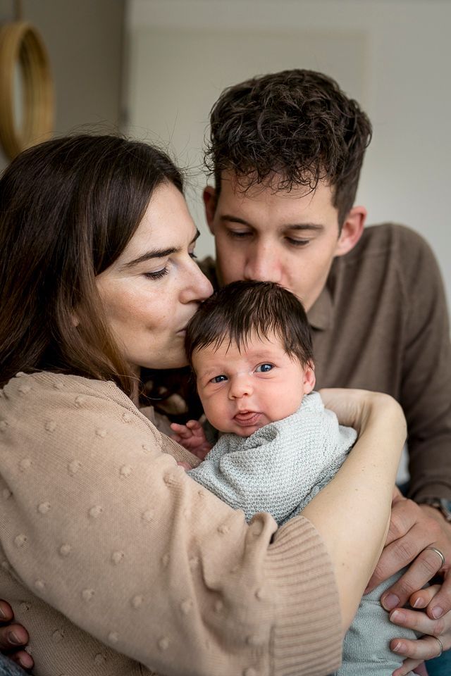 Ouders kussen het hoofdje van hun pasgeboren baby. Moeder houdt de baby vast, vader buigt zich voorover. Binnen, zacht licht.