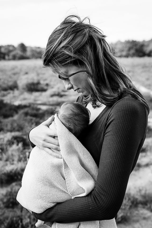 Vrouw met een ingewikkelde baby in haar armen, kijkt naar beneden. Buiten, wazig landschap op de achtergrond. Zwart-wit.