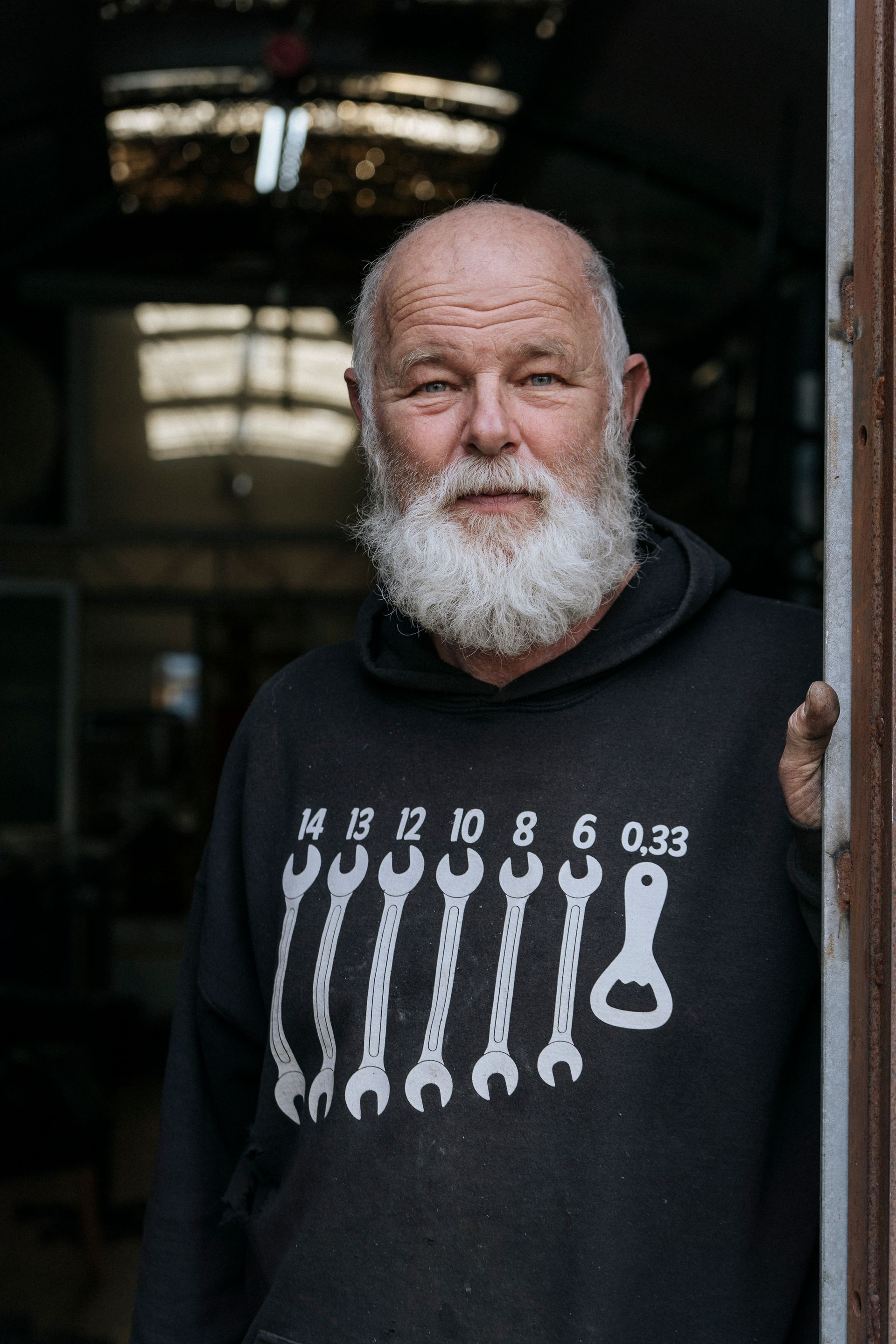 Een man met een witte baard en een zwarte hoodie met moersleutelmotief staat in een deuropening.