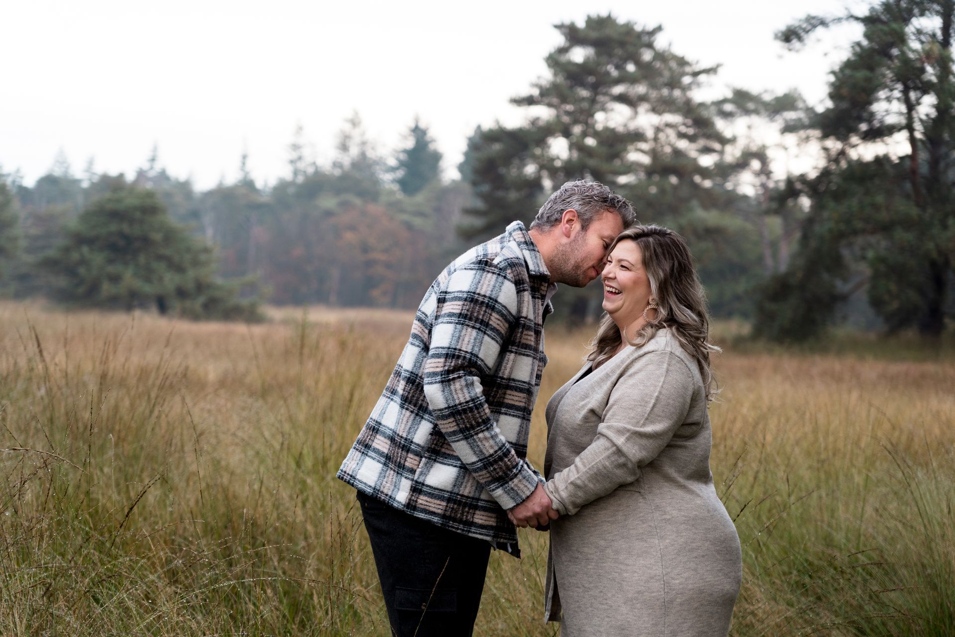 Een stel loopt hand in hand in een grasveld. De man kust de vrouw op haar voorhoofd; beiden glimlachen. Op de achtergrond zijn bomen te zien.