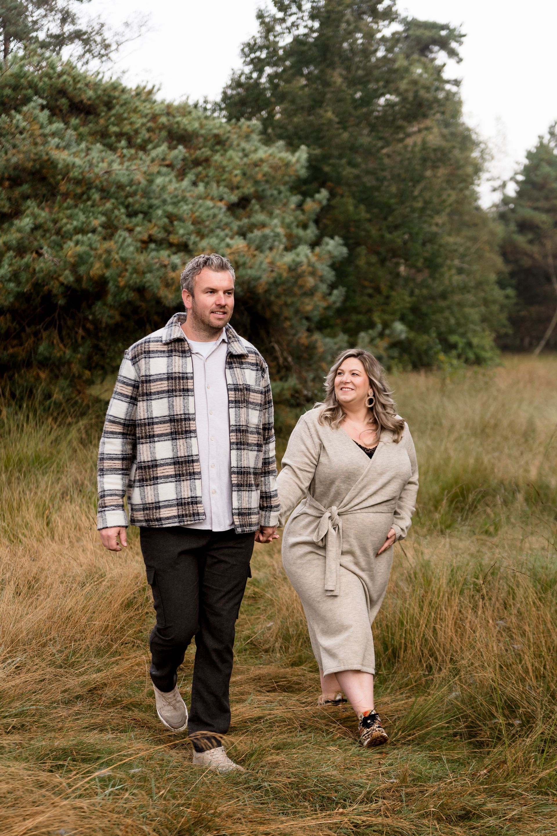 Een stel loopt hand in hand door een veld; de man draagt ​​een geruit overhemd, de vrouw een beige jurk, met bomen op de achtergrond.