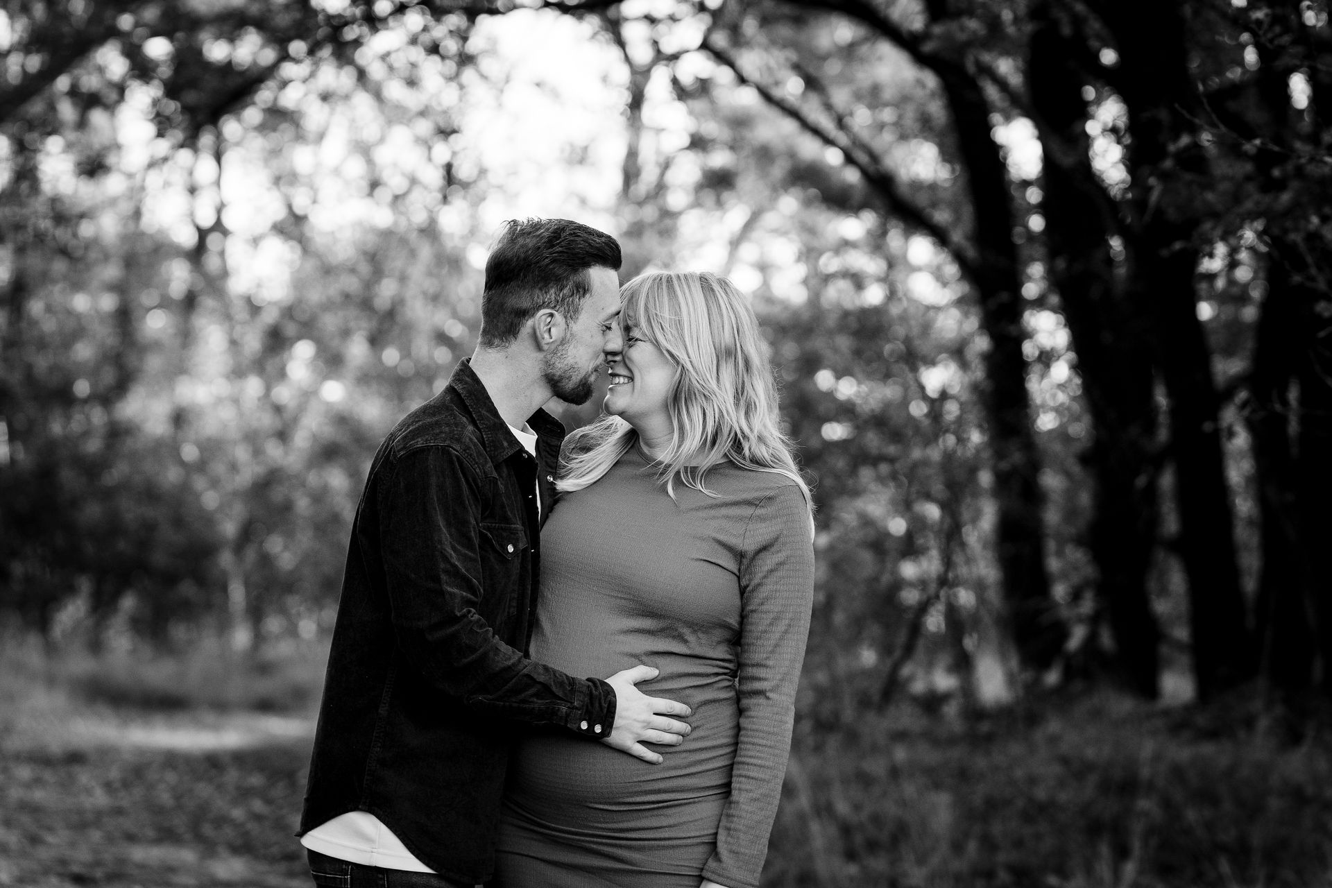 Een stel omhelst elkaar in een bos; de man kust de vrouw op haar wang, en ze is zwanger. Zwart-wit.