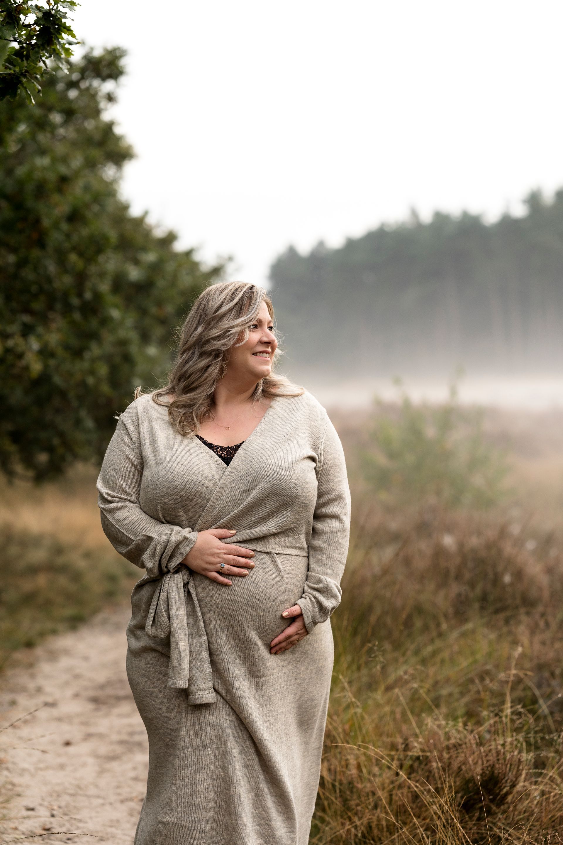 Een zwangere vrouw in een lange beige trui, met haar hand op haar buik, lachend, staat op een pad in een mistig bos.