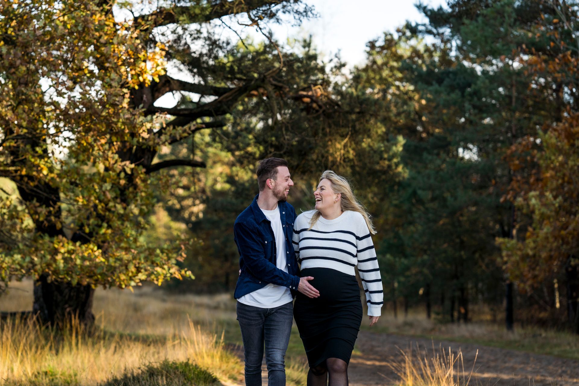 Een man en een zwangere vrouw lopen over een pad in een bosrijk gebied. De man glimlacht en raakt de buik van de vrouw aan.
