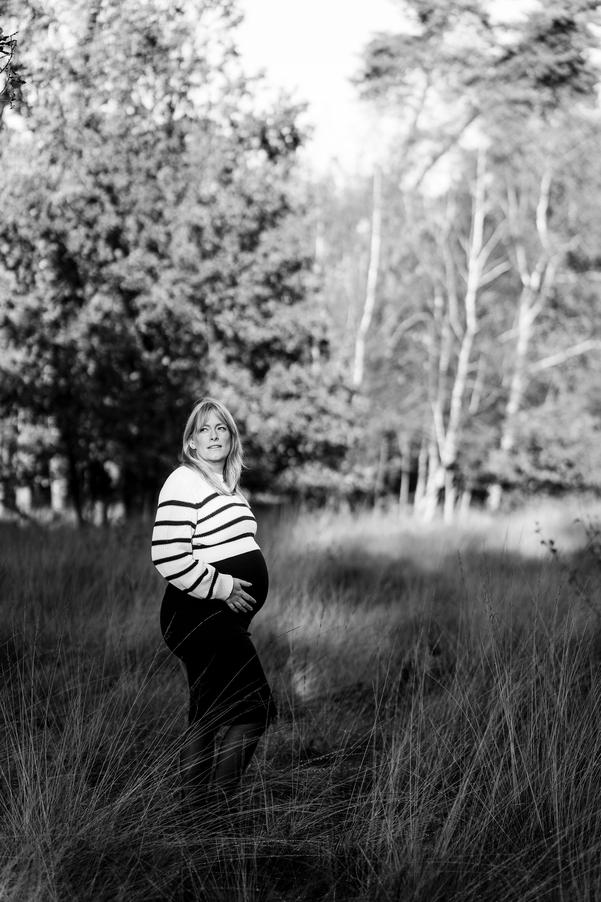 Een zwangere vrouw staat in een grasveld, met haar handen op haar buik, en draagt ​​een gestreepte trui.