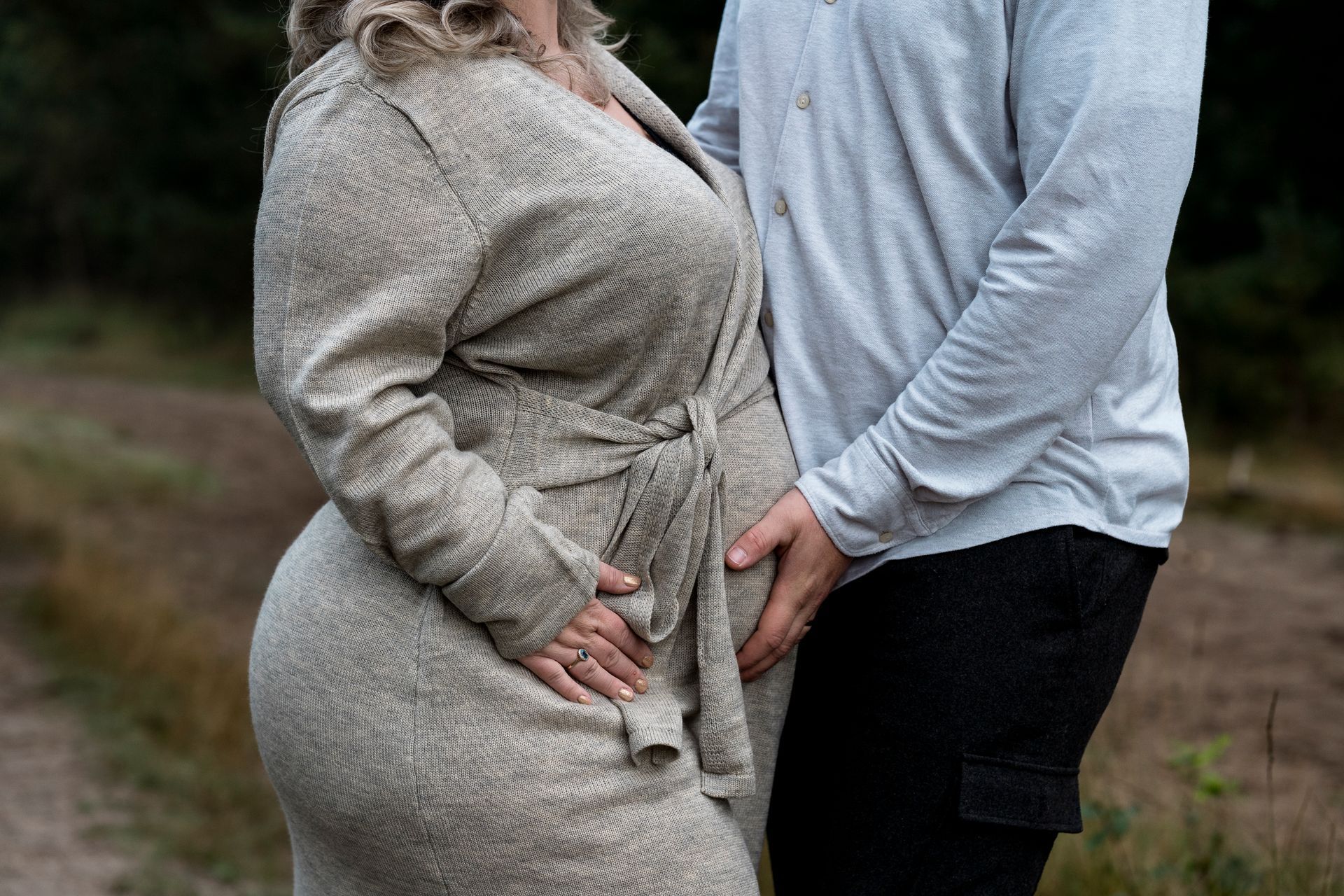 Zwangere vrouw in beige badjas met de hand van haar partner op haar buik, buiten.