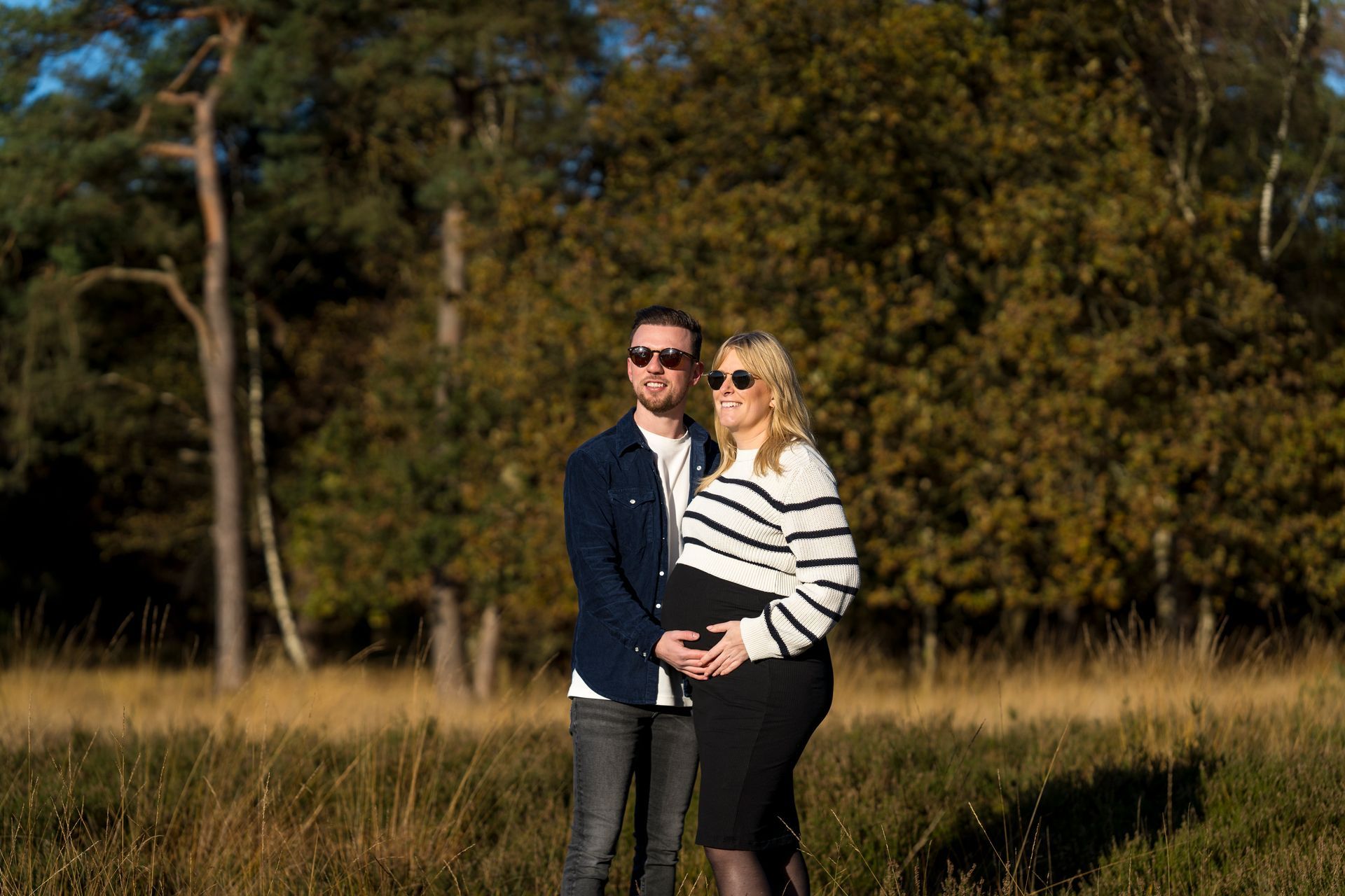 Een stel met zonnebrillen poseert buiten; een vrouw houdt haar zwangere buik vast en staat in een veld.