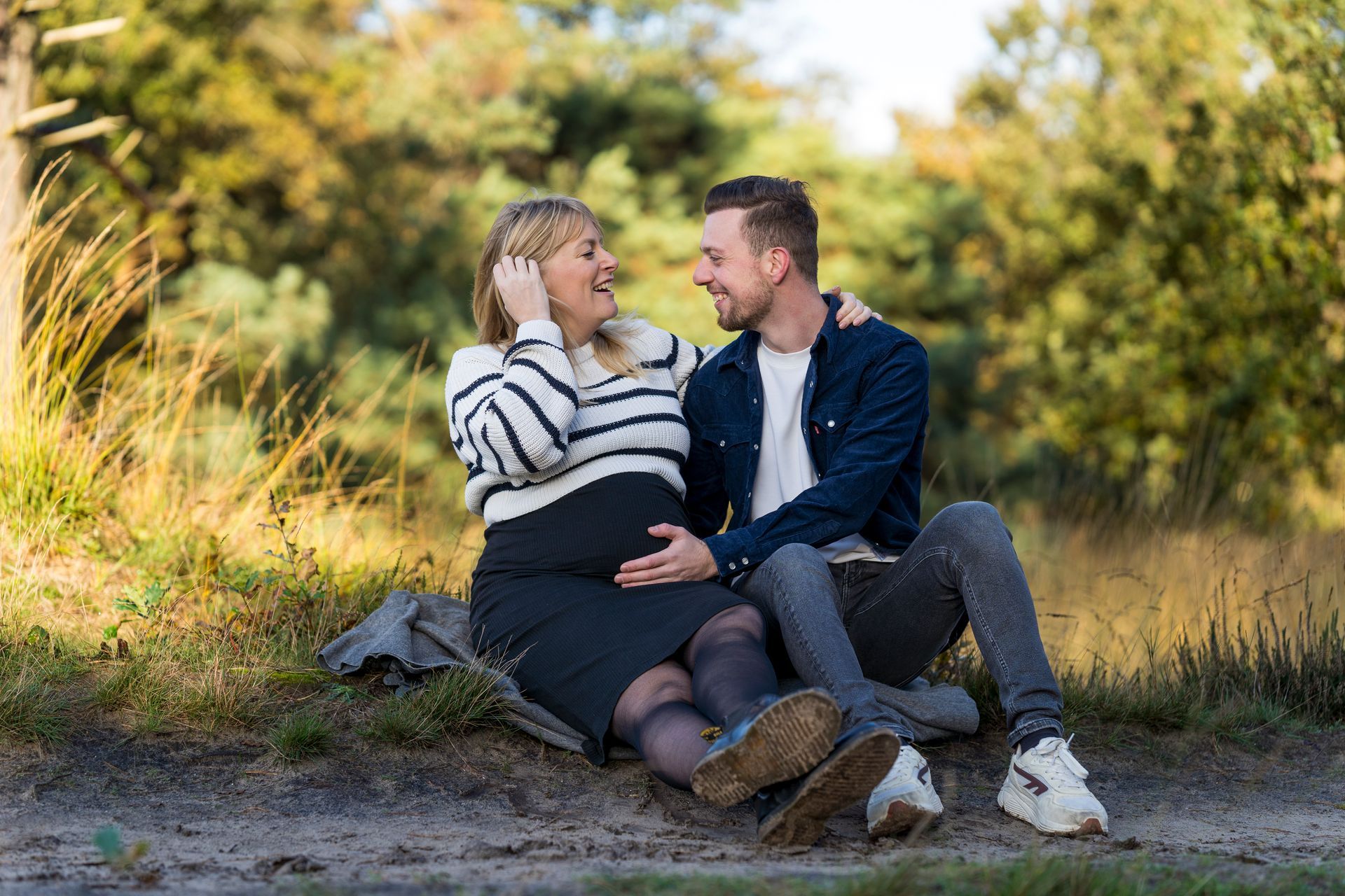 Een zwanger stel zit buiten, lachend en elkaar omhelzend. De vrouw draagt ​​een gestreepte trui, de man een spijkerjasje. Op de achtergrond is een bos te zien.
