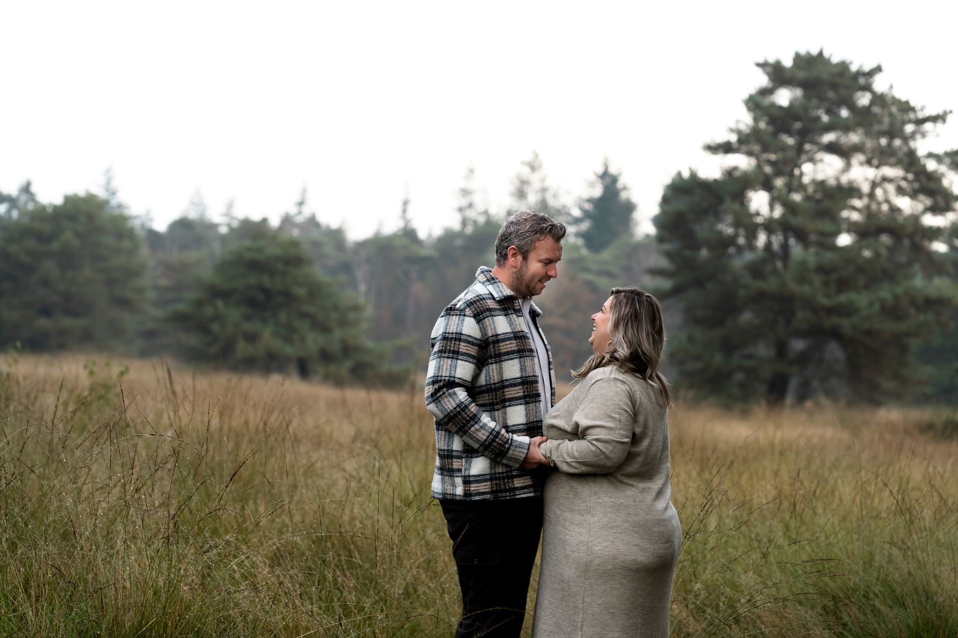 Een stel loopt hand in hand en kijkt elkaar aan in een veld met hoog gras. Bewolkte lucht, bomen op de achtergrond.