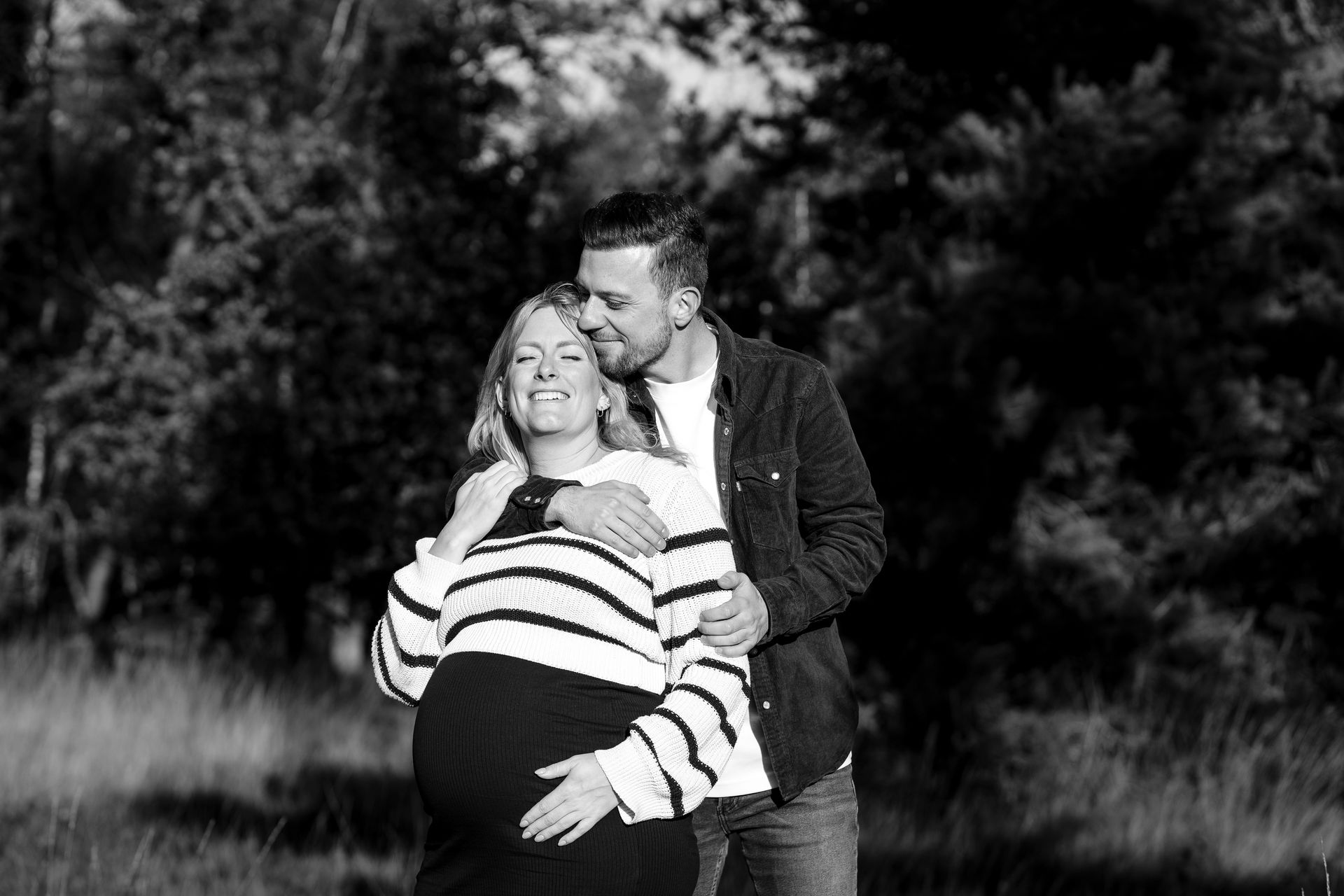Een zwangere vrouw en een man omhelzen elkaar in de buitenlucht en glimlachen. De man heeft een arm om de schouder van de vrouw geslagen; ze staan ​​voor een groep bomen.