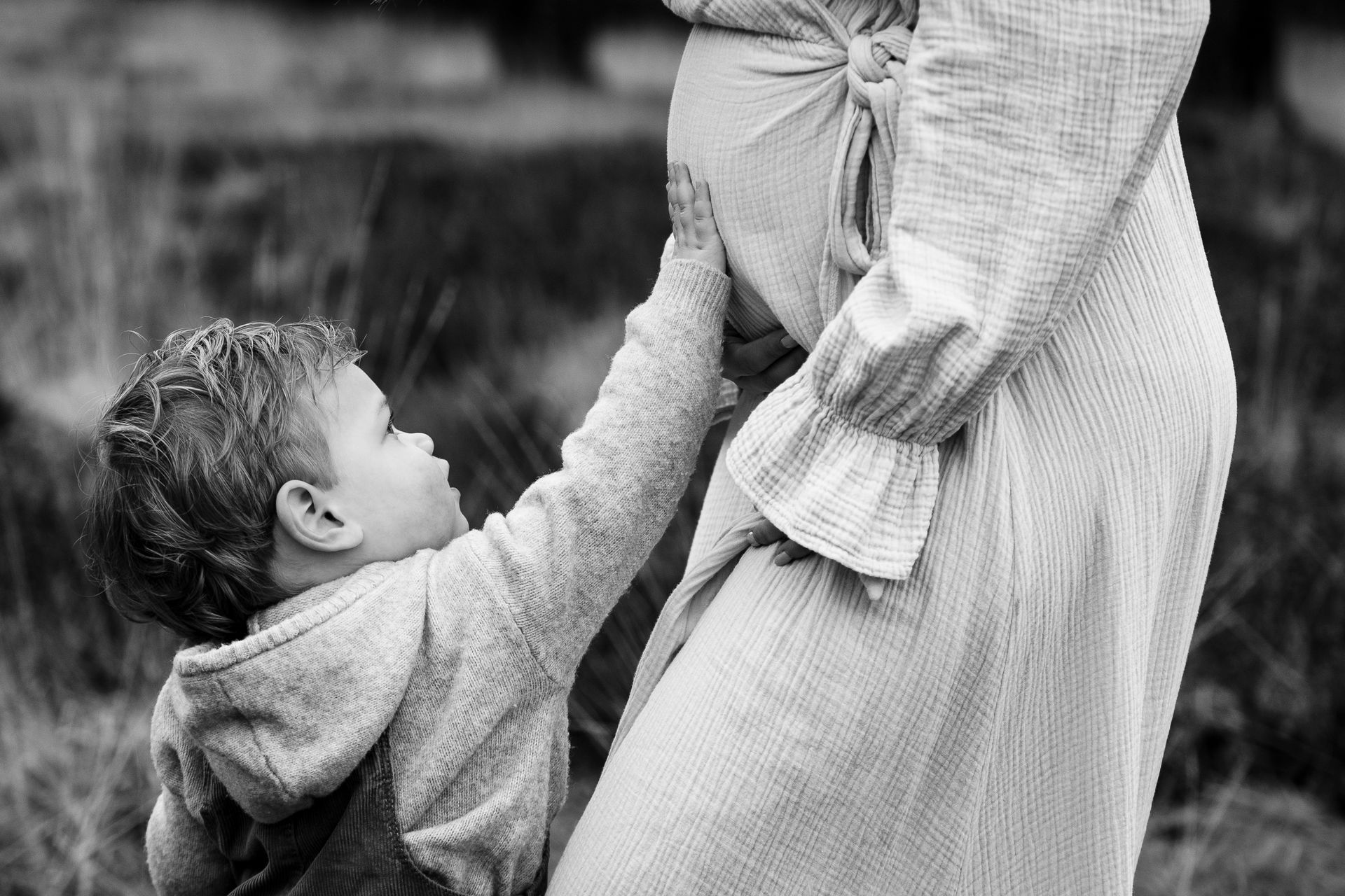 Jongen raakt zwangere buik aan in de buitenlucht. Jurk van een vrouw met strikceintuur. Zwart-wit.