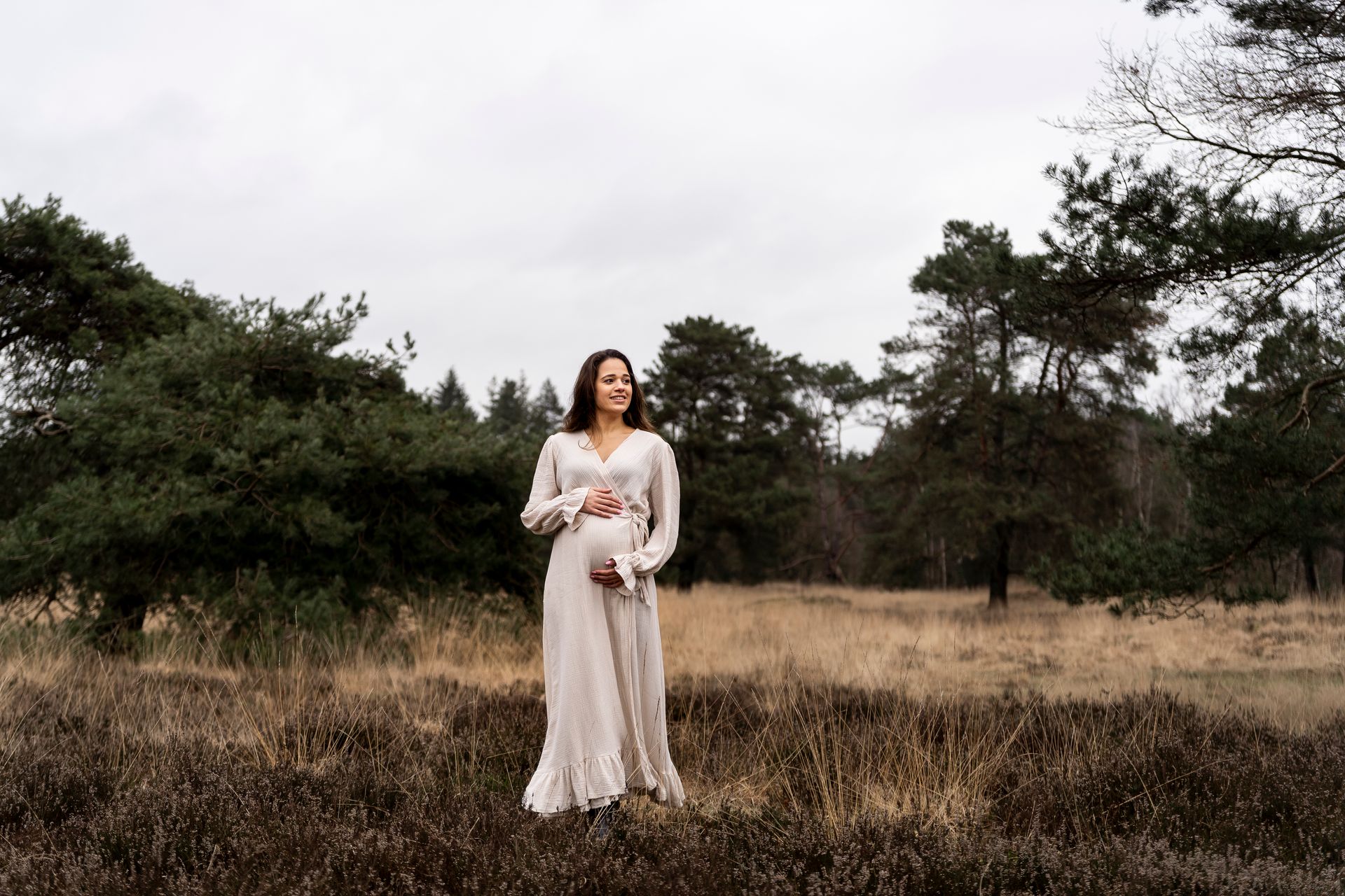 Een zwangere vrouw in een beige jurk staat in een veld met bomen.