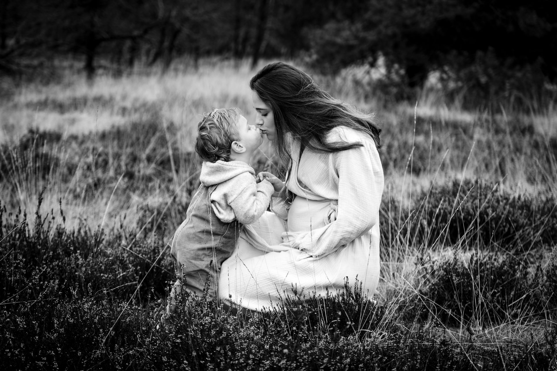 Een zwangere vrouw met haar kind in een veld, waarbij het kind de wang van de vrouw aanraakt. Zwart-wit.