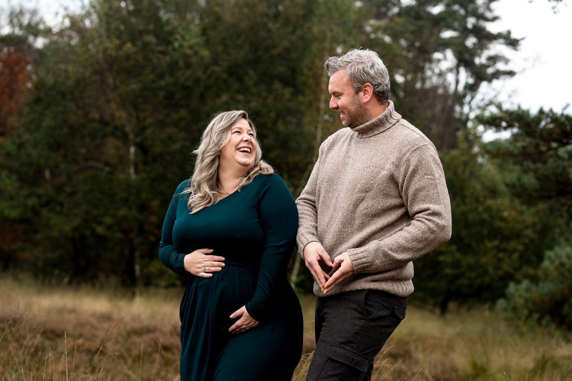 Een lachend stel in de buitenlucht. Een zwangere vrouw in een turquoise jurk; een man die met zijn handen een hartje vormt.