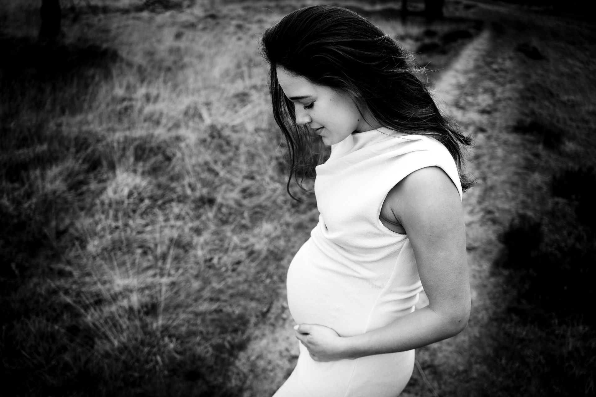 Zwangere vrouw in een mouwloze jurk, die haar buik vasthoudt en naar beneden kijkt. Buiten op een pad, zwart-wit.