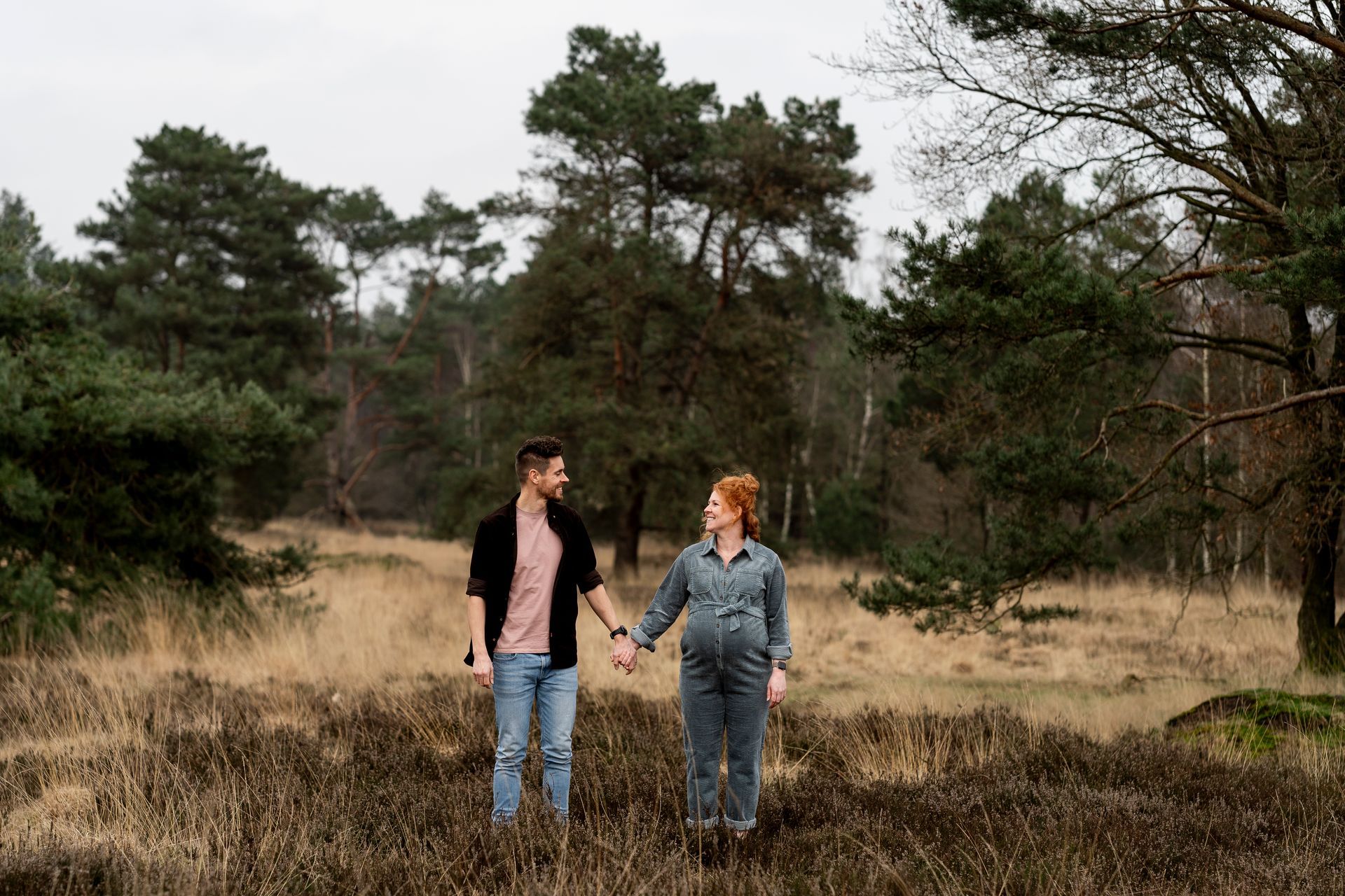 Een stel, hand in hand, verwacht een kindje. Ze staan ​​in een veld met bomen op de achtergrond. De lucht is bewolkt.