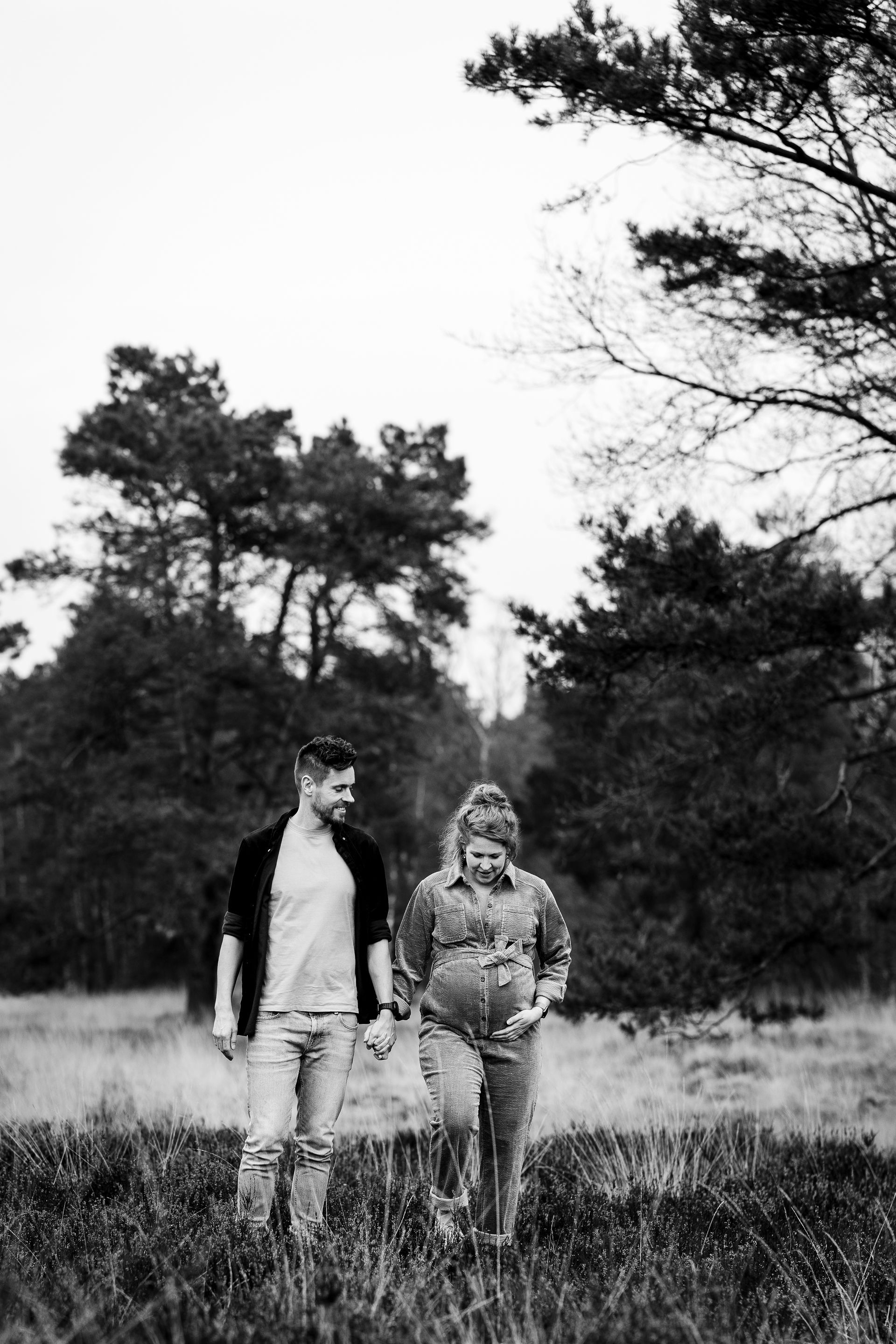 Een stel loopt hand in hand in een veld; de vrouw is zwanger en de man kijkt naar haar. Zwart-witfoto.