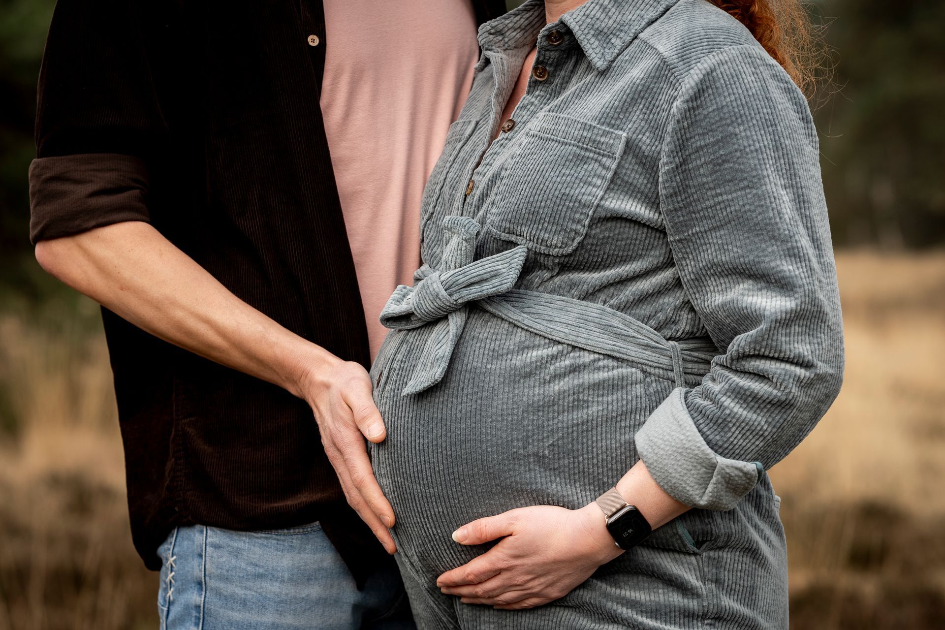 Een man en een zwangere vrouw houden elkaars hand vast boven een babybuik; buiten, neutrale tinten.