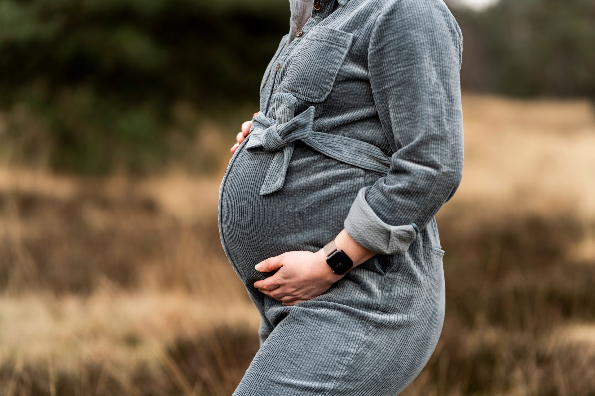 Zwangere vrouw in grijze overall, handen op de buik, buiten.