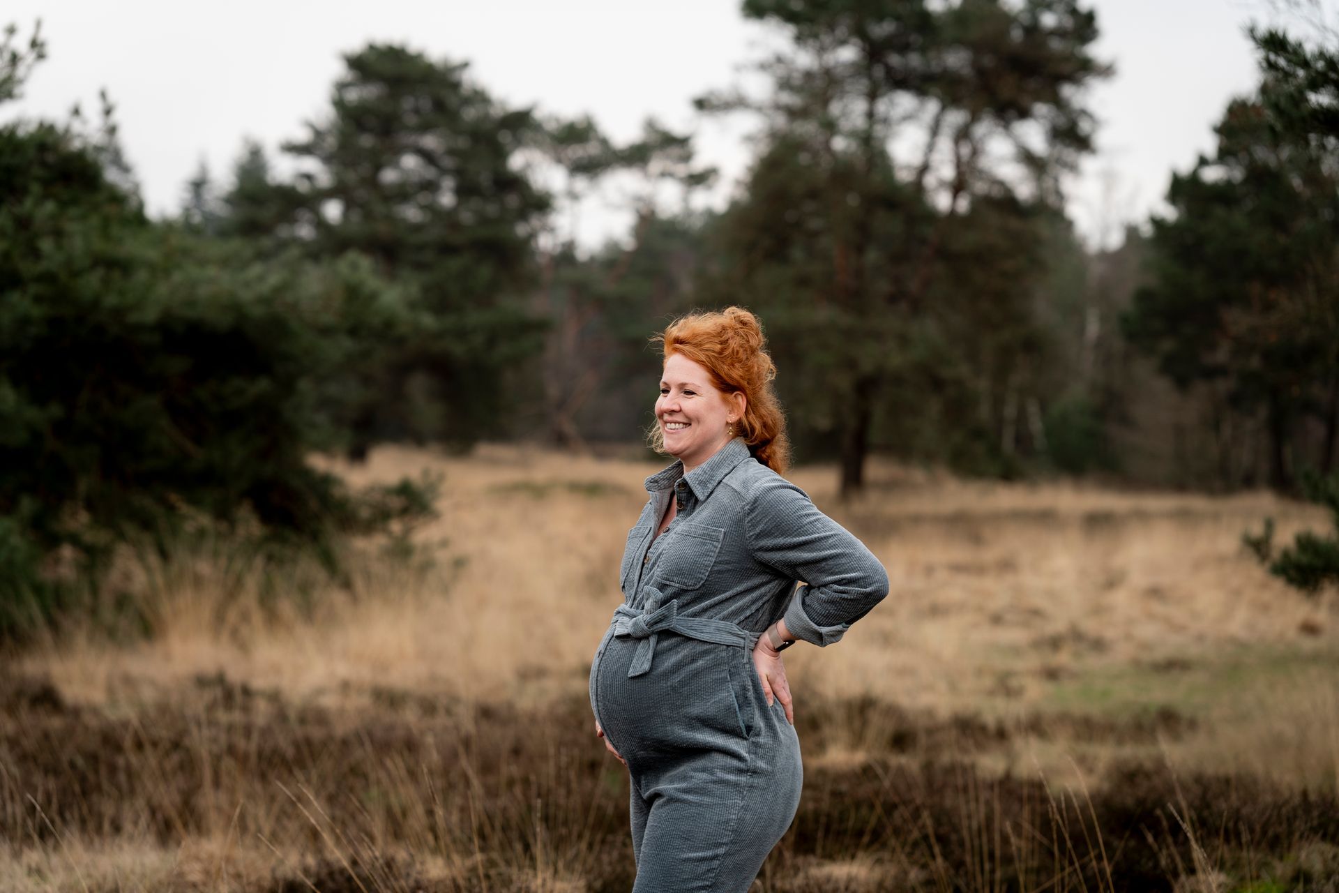 Een zwangere vrouw lacht, zet haar handen in haar zij en draagt ​​een spijkeroverall in een veld met bomen.