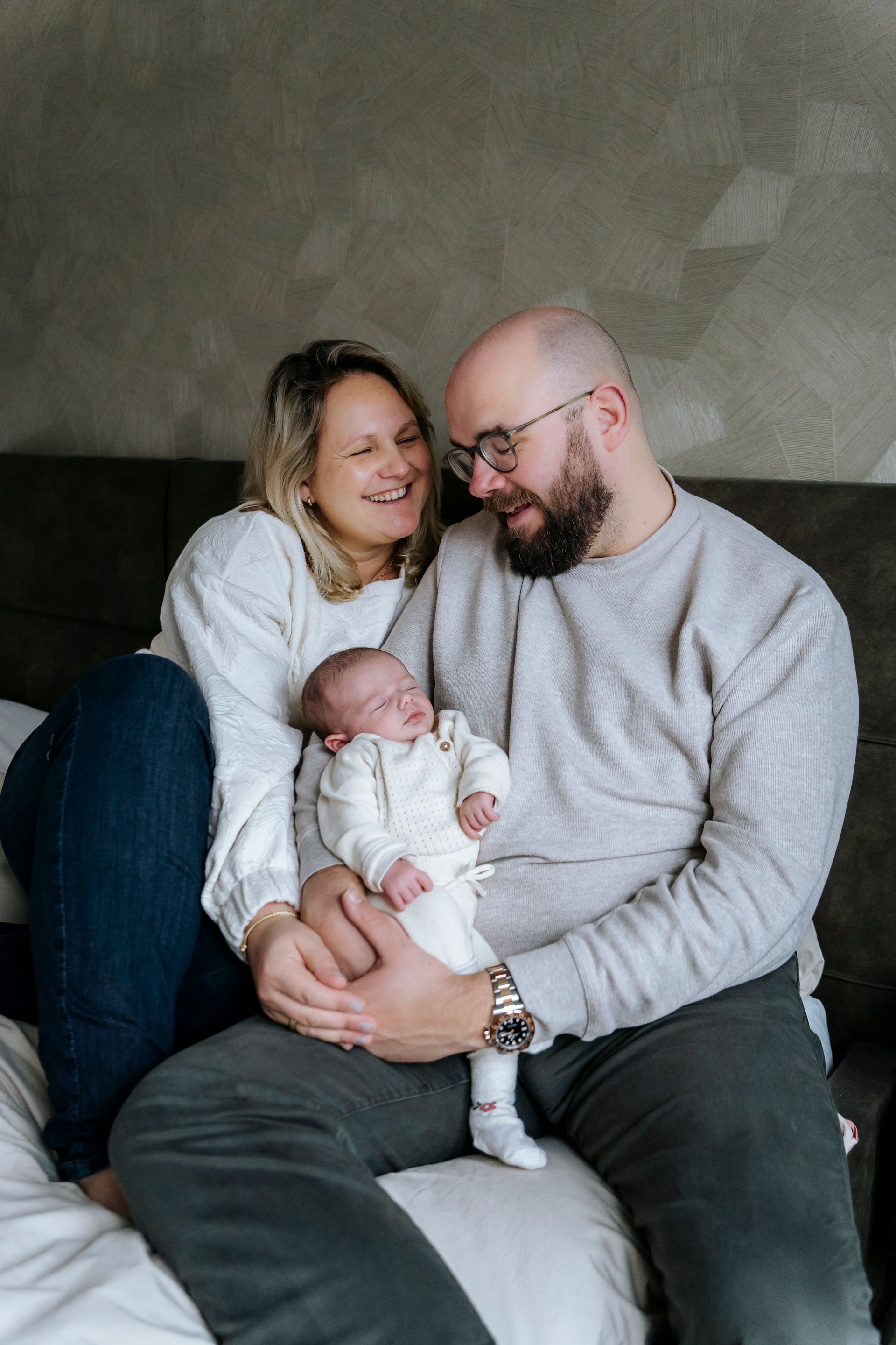 Ouders houden een pasgeboren baby vast op een bed. Moeder glimlacht, vader kijkt naar beneden; zachte, neutrale kleuren.