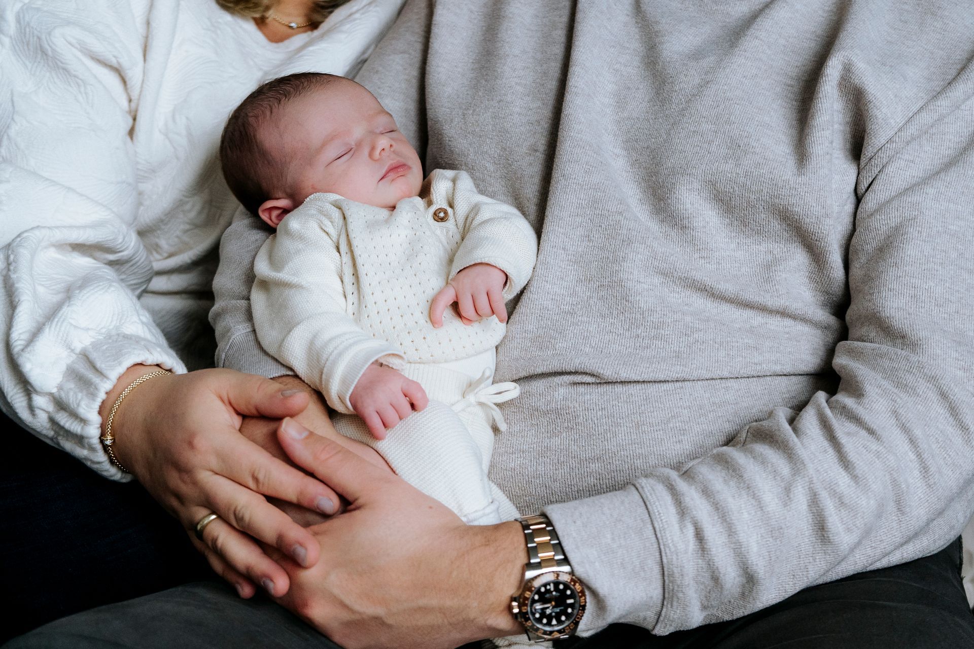 Pasgeboren baby slaapt in de armen van de ouders; de baby draagt ​​een wit gebreid pakje.