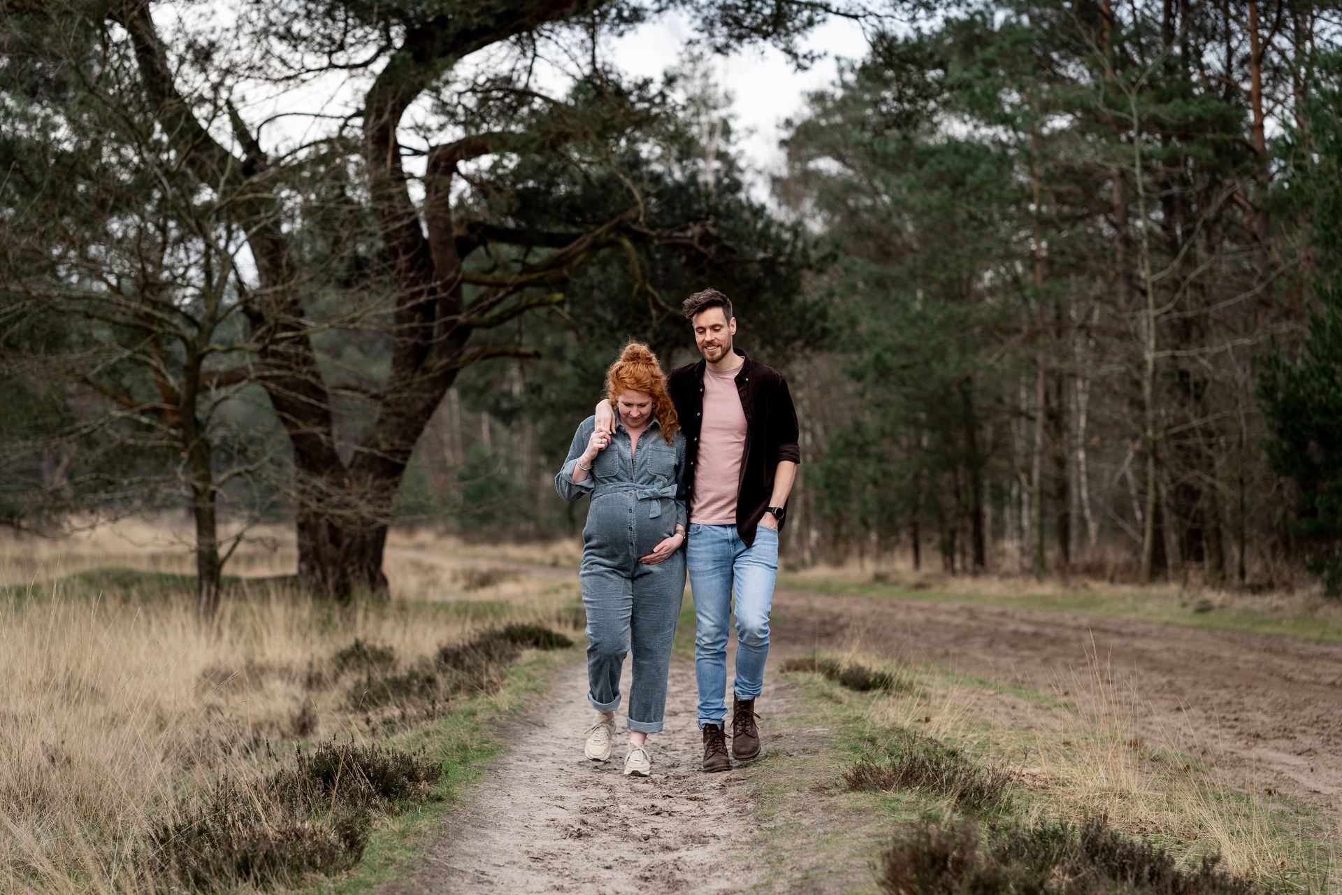 Een zwangere vrouw en haar partner lopen over een pad in een bosrijk gebied. De zwangere vrouw draagt ​​een spijkeroverall en haar partner een overhemd en een spijkerbroek.