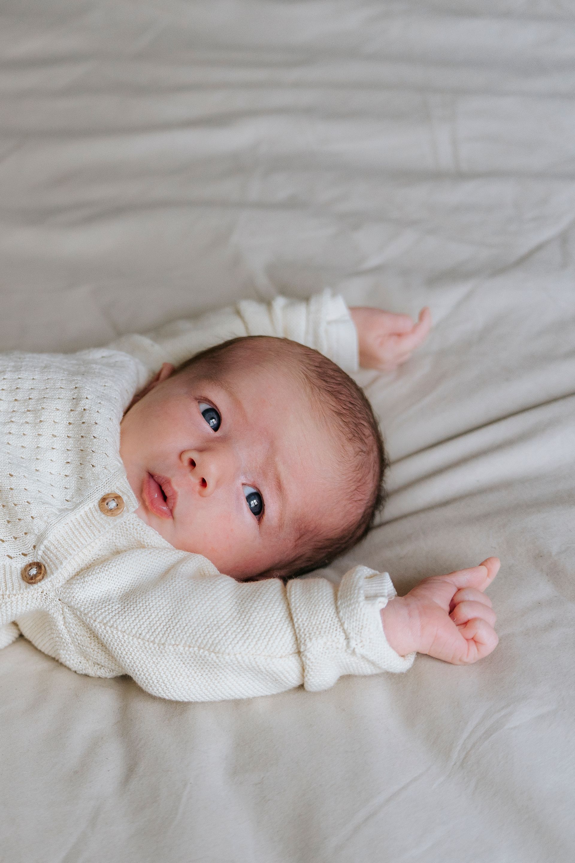 Baby in een wit truitje ligt op een lichtgekleurde ondergrond, met uitgestrekte armen, en kijkt naar de camera.