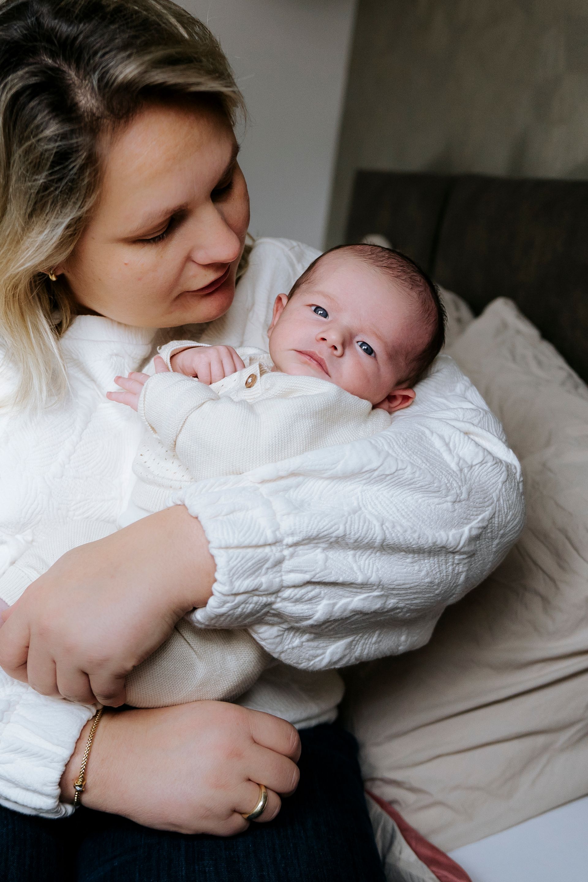 Vrouw in witte trui met een baby op haar arm; beiden kijken naar de camera.